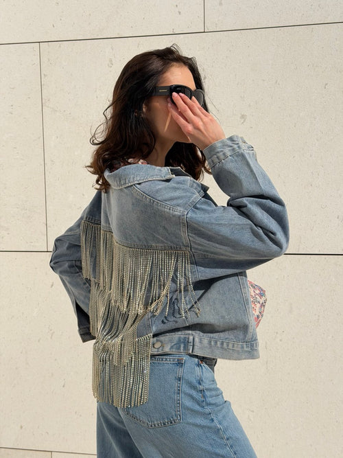 Person wearing a denim jacket with fringe details against a light-colored wall.