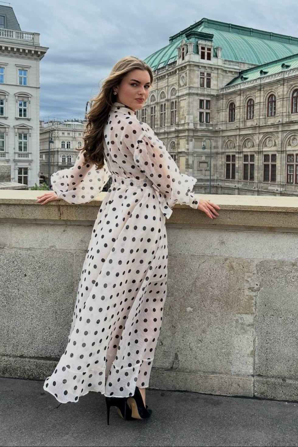 Woman in a white dress with black polka dots standing on a stone wall with a building in the background.